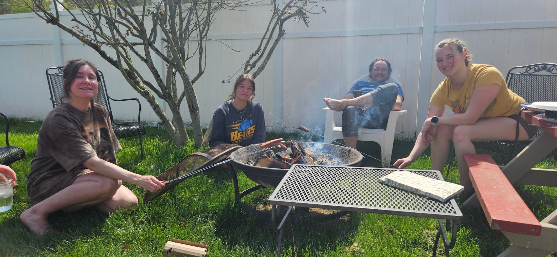 Family sitting around a backyard fire pit roasting hot dogs, with Tobe barefoot in a lawn chair.