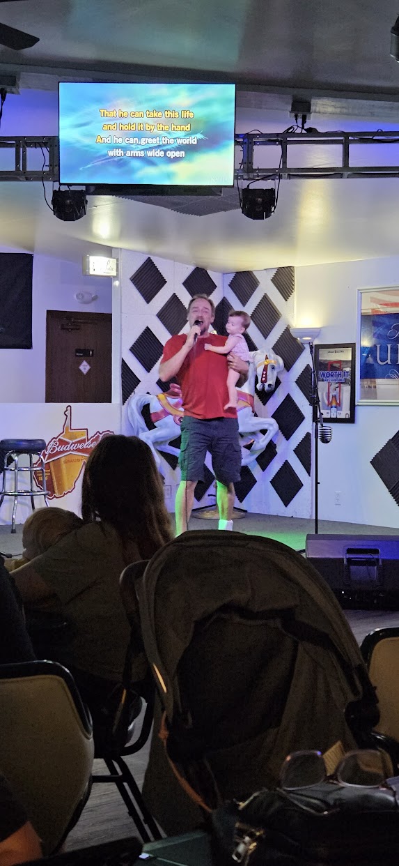 Tobe singing karaoke on stage while holding his baby granddaughter, with the lyrics on a screen above.